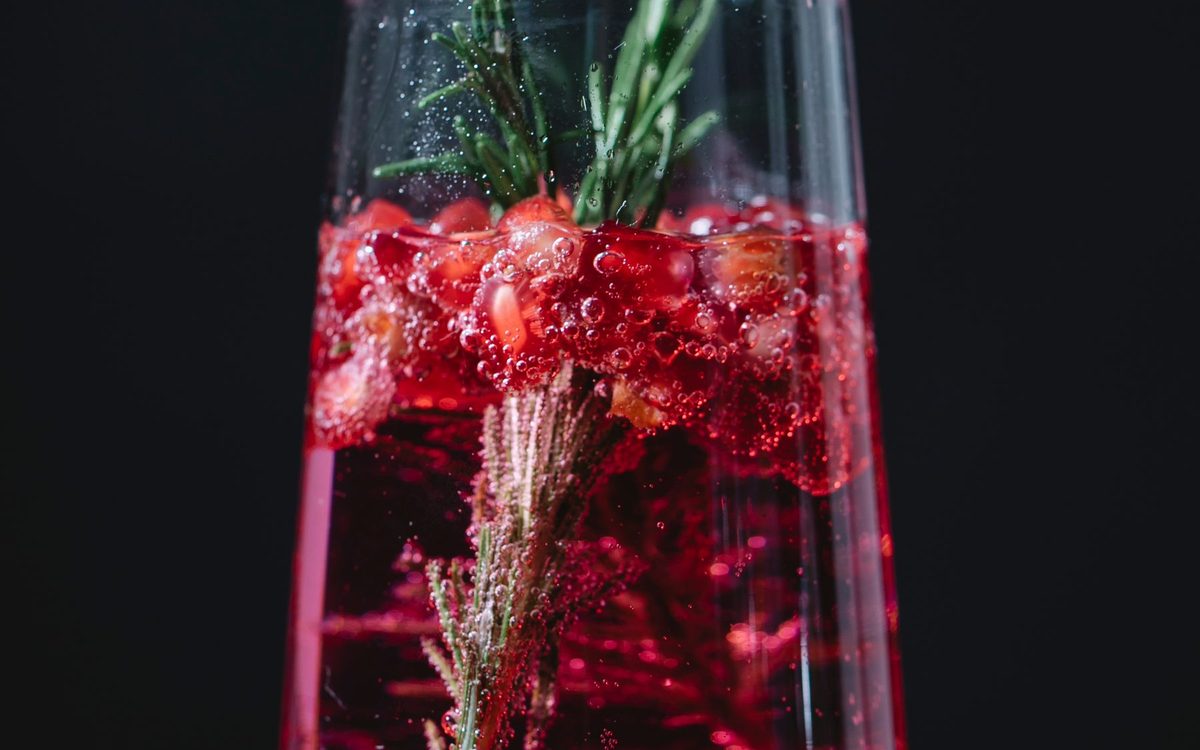 Tall red cocktail with pomegranate seeds and a sprig of rosemary