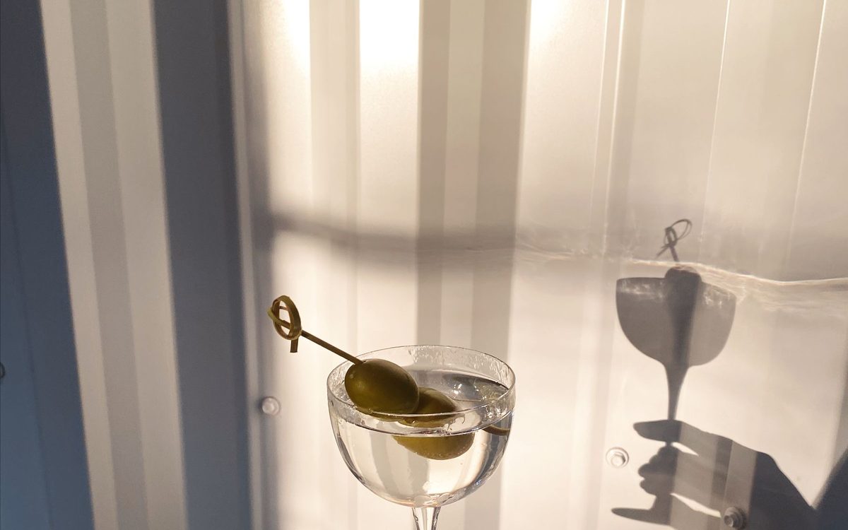 A hand holding a martini glass with olives against a pale curtain